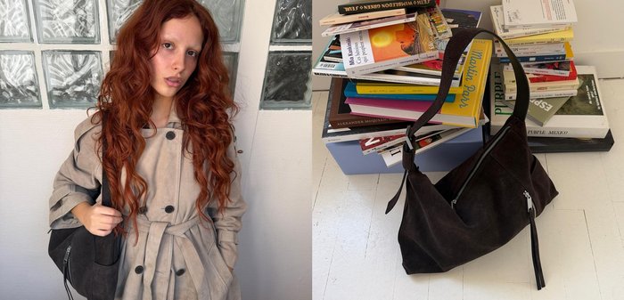 Jeune femme aux longs cheveux roux bouclés portant un trench beige, tenant un sac à bandoulière noir, debout contre un mur en blocs de verre.