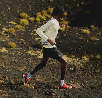 Coureur portant un maillot à manches longues blanc et des chaussures rouges, courant sur un sentier rocheux à végétation clairsemée en plein air dans un cadre naturel.