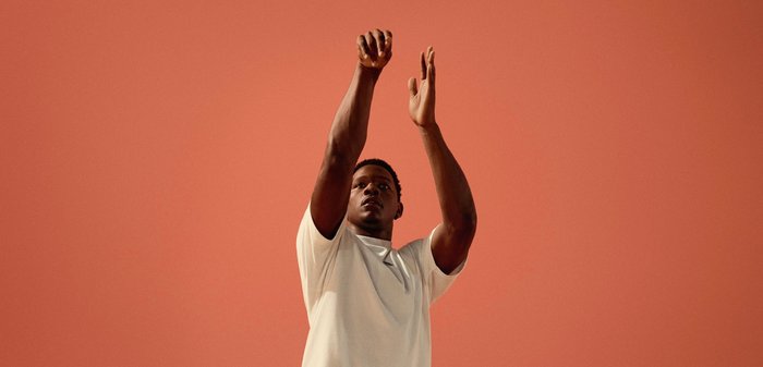 Chemise blanche à manches courtes portée par une personne levant le bras sur un fond corail uni. Design épuré, texture douce, sans motifs visibles.