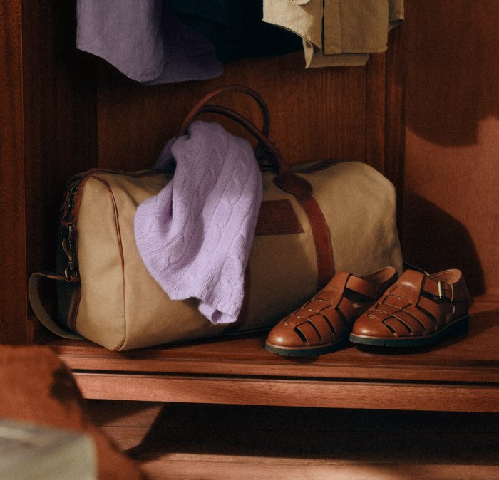 Sandali in pelle e un maglione lavorato a maglia color lavanda posati su una borsa da viaggio beige dentro un armadio di legno.