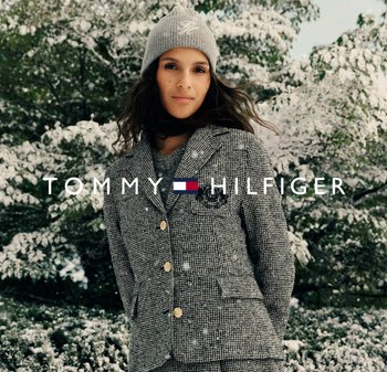 Sakko mit Hahnentritt-Muster und goldenen Knöpfen, kombiniert mit einer hellgrauen gestrickten Mütze. Schneebedeckter Hintergrund verstärkt winterliche Atmosphäre.