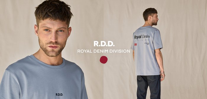 Man draagt een lichtgrijs T-shirt van Royal Denim Division en een donkere jeans, staat zijwaarts tegen een effen beige achtergrond.