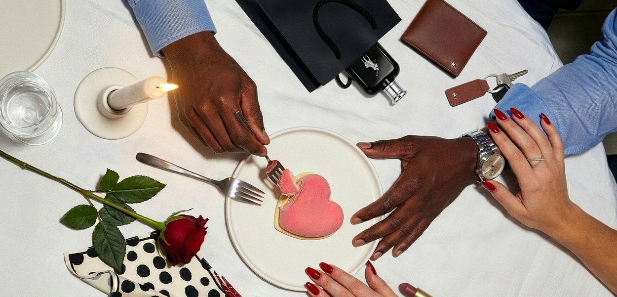 Manos compartiendo un postre en forma de corazón en una mesa iluminada por velas, con una rosa, cartera, llaves y lápiz labial rojo visibles, sugiriendo un ambiente romántico.