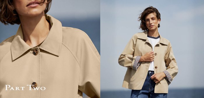 Femme portant une veste beige à boutons avec des poignets à carreaux, debout dehors sous un ciel nuageux avec les cheveux soufflés par le vent.