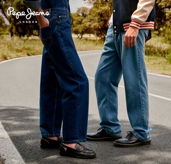 Dos personas vestidas con prendas de mezclilla están de pie en una carretera. Una lleva jeans azul oscuro, mientras que la otra usa jeans azul claro y zapatos negros.