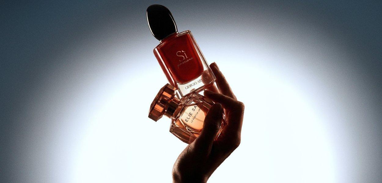 Hand, die zwei Parfümflaschen hält, eine rote mit der Aufschrift "Si" von Giorgio Armani, die andere kleiner, beleuchtet vor einem grauen Verlaufs-Hintergrund.