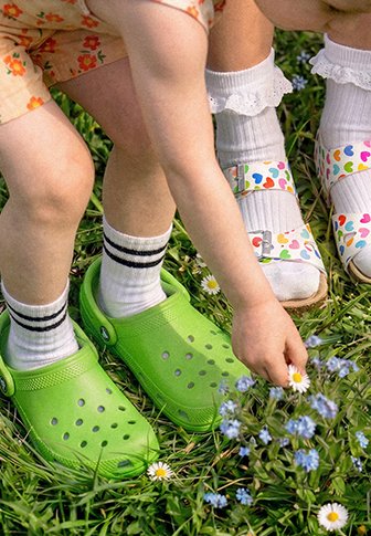 Twee kinderen plukken madeliefjes in het gras; de een draagt groene klompen en gestreepte sokken, de ander witte sandalen met hartjespatroon en gerimpelde sokken.