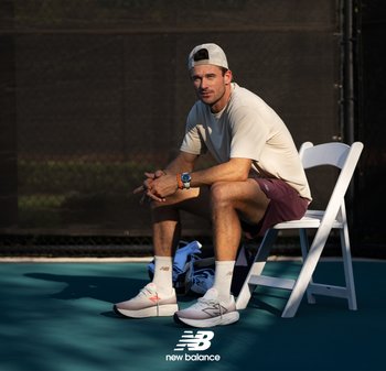 Des chaussures de sport New Balance en rose clair avec des accents plus foncés, un t-shirt beige et un short bordeaux. Une chaise pliable blanche, en arrière-plan un court de tennis bleu.