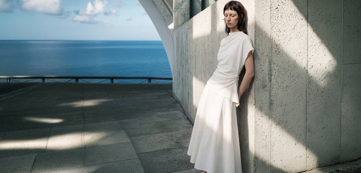 Una mujer con un vestido blanco está de pie con los ojos cerrados contra una pared de cemento iluminada por el sol cerca de una vista al océano bajo un cielo parcialmente nublado.