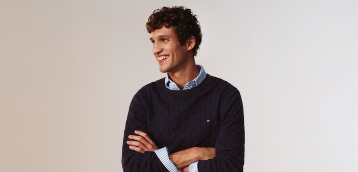 Ung mand med krøllet hår iført en navyblå sweater og en blåstribet skjorte, smilende, med armene over kors, kigger til siden mod en enkel baggrund.