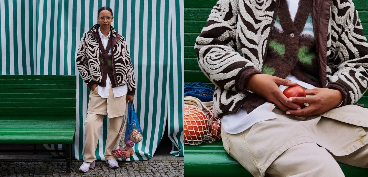 Brun og hvit mønstret cardigan, kremfarget skjorte og bukser, rosa sneakers. Holder et rødt eple, med en grønn benk og stripete bakgrunn.