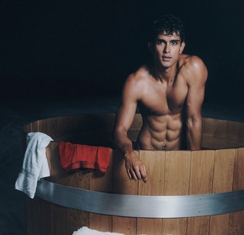 Träbadkar med ett metallband. En person med fuktigt hår lutar sig ut, med en vit handduk och röda shorts hängande över kanten. Mörk bakgrund.