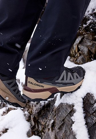 Ο πεζοπόρος φοράει μποτάκια Salomon και σκαρφαλώνει σε χιονισμένο και βραχώδες έδαφος, με μαύρα παντελόνια ορατά πάνω από τα μποτάκια.