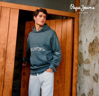 Giovane uomo con felpa blu e pantaloni bianchi in piedi davanti a una porta di legno, che guarda di lato, con sfondo di muro in pietra e logo Pepe Jeans London.