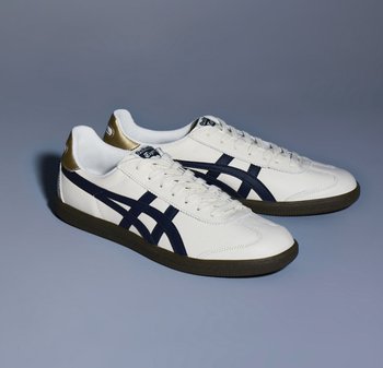 Baskets de sport blanches avec des rayures bleu marine et un accent doré à l'arrière. Elles possèdent une semelle en caoutchouc et un design à lacets.