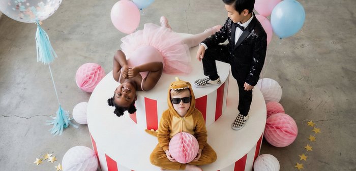 Trois enfants en costumes posent sur une plateforme à étages avec des ballons et des décorations en papier lors d'une fête intérieure festive.