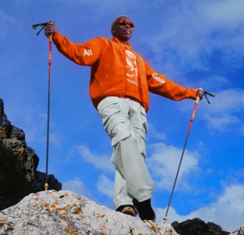 Person trägt eine orange Jacke und Cargohosen und wandert mit Trekkingstöcken auf felsigem Gelände unter blauem Himmel.