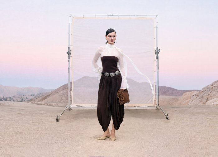 Femme debout sur un sol désertique portant un chemisier blanc transparent à col haut et une combinaison noire bustier, tenant une pochette marron, avec des montagnes en arrière-plan.