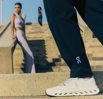 Chaussons sportifs blancs avec une semelle texturée, associés à un pantalon sombre et ample. Deux personnes en tenue de sport sur des escaliers en arrière-plan.