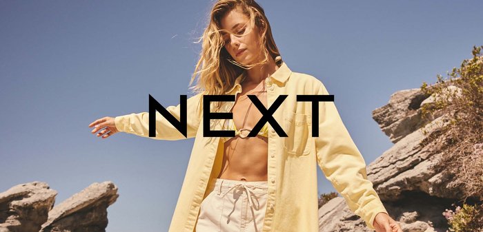Femme portant une chemise jaune par-dessus un haut de bikini et un short blanc, debout, les bras étendus, sur une plage rocheuse sous un ciel bleu clair.