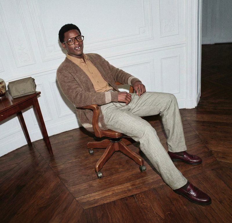 Férfi szemüvegben, bézs ingben, barna kardigánban és világos nadrágban, egy sötét parkettás padlón álló forgószéken ül.