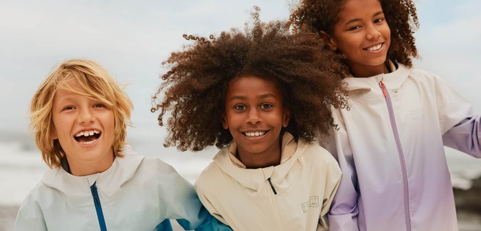 Three smiling children outdoors wearing light-colored jackets, with one child linking arms with another, against a cloudy sky background.