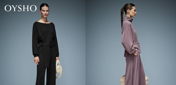 Frau mit langen geflochtenen Haaren, die einen übergroßen Trainingsanzug in Mauve trägt, seitlich stehend mit den Händen in den Taschen vor einem schlichten grauen Hintergrund.