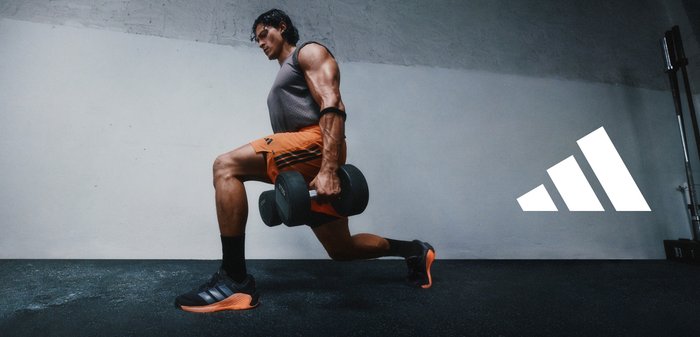 Black dumbbells held by a person wearing a grey vest and orange shorts, performing a lunge on a dark surface with textured flooring.
