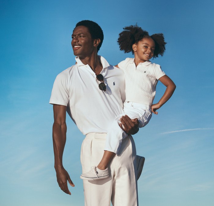 Hombre con polo blanco y pantalones sostiene a una niña sonriente con ropa blanca contra un cielo azul despejado al aire libre.