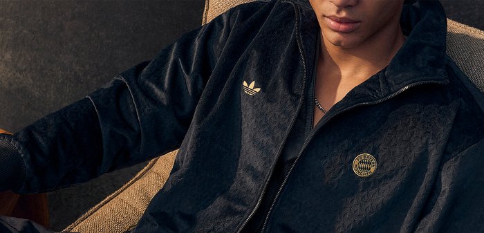 Jeune personne portant une veste de survêtement Adidas noire avec le logo du FC Bayern Munich, assise sur une chaise beige texturée.