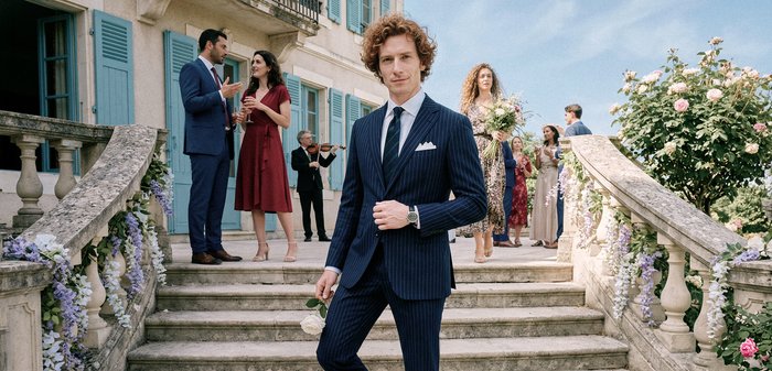 Άνδρας με μπλε κοστούμι με ρίγες κρατάει ένα λευκό τριαντάφυλλο σε διακοσμημένα πέτρινα σκαλοπάτια, με ομάδες ανθρώπων να κοινωνικοποιούνται με κομψή ενδυμασία.