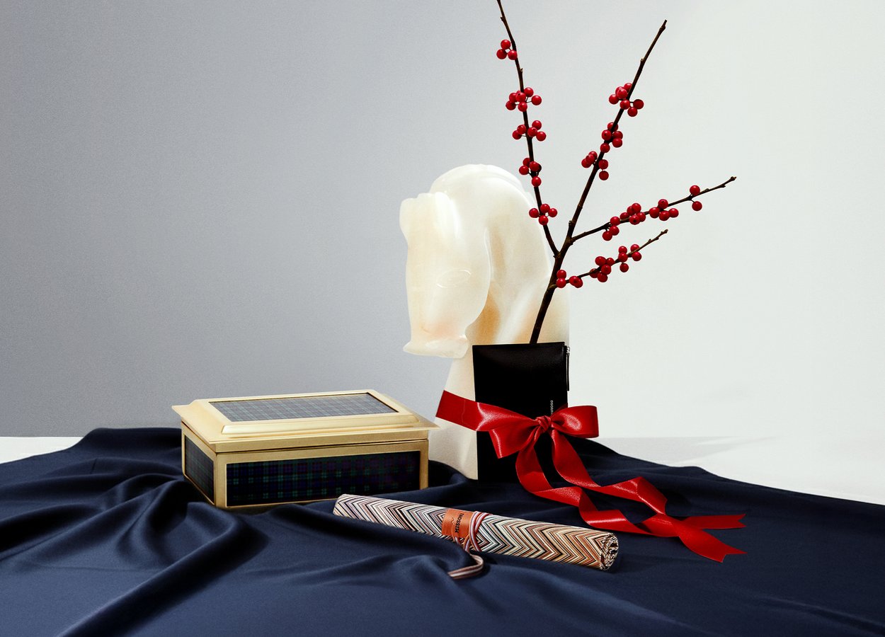 Vaza v obliki belega konjskega glave drži veje z rdečimi jagodami. Poleg je zlata škatla s karo pokrovom in zvit darilni papir s cikcak vzorcem na temno modri tkanini.