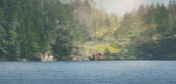 Cabane en bois rouge avec un toit en pente, située sur un rivage rocheux, entourée d'arbres verts luxuriants, face à un lac bleu calme.