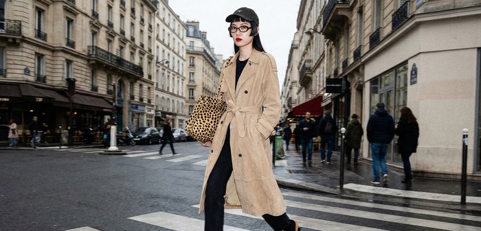 Femme portant un trench beige et une casquette noire traversant une rue de la ville, tenant un sac fourre-tout à imprimé léopard, avec des bâtiments et des piétons en arrière-plan.