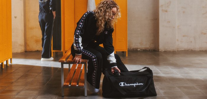 Black gym bag with white logo, made of fabric. Model in athletic wear with logo accents, seated on a wooden bench in a locker room.