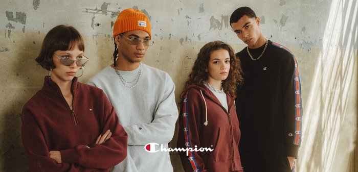 Quatre mannequins portant des vêtements décontractés de la marque Champion, debout contre un mur gris texturé avec des expressions neutres et un éclairage minimal.