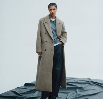 Brauner Wollmischmantel mit breiten Revers und doppelt geknöpft, kombiniert mit einem grauen Poloshirt und schwarzen Hosen auf einer strukturierten dunklen Oberfläche.