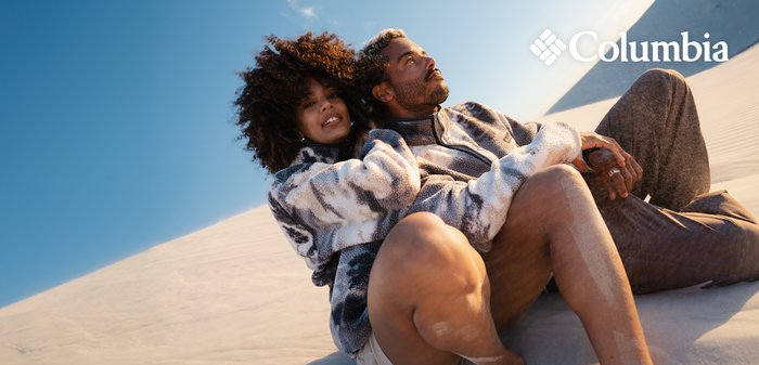 Couple in patterned fleece jackets sitting on sand dunes under clear sky, relaxed and close, Columbia logo in top right corner.