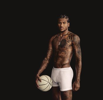 Boxer Calvin Klein bianchi indossati da una persona tatuata che tiene un pallone da basket, su sfondo nero.