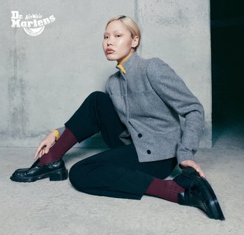 Chaqueta de tweed gris con cuello alto, combinada con pantalones negros y zapatos negros gruesos, complementada con calcetines burdeos. Fondo de concreto.