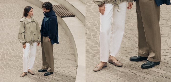 Twee volwassenen staan op een met stenen geplaveide ondergrond. De één draagt een beige jasje, witte broek en lichtbruine flats; de ander heeft een marineblauwe sjaal om, een bruine broek en zwarte schoenen aan.