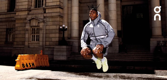 Athlete with dreadlocks jumping in front of stone columns, wearing a gray jacket, black shorts, white socks, and neon yellow running shoes.