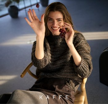 Gebreide trui in donkere tinten met geribbelde textuur, gedragen met hoge, donkere broeken. Houdt paarse druiven vast, zittend op een houten stoel.