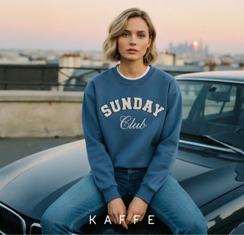 Blauwe sweatshirt met "SUNDAY Club" geborduurd in crème, gedragen met blauwe jeans, zittend op een zwarte auto, tegen een zonsondergang stadslandschap.