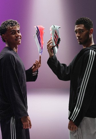 Zwei Männer stehen sich gegenüber und halten schwebende Fußballschuhe, einer pink und der andere silber, vor einem lila Farbverlauf-Hintergrund.