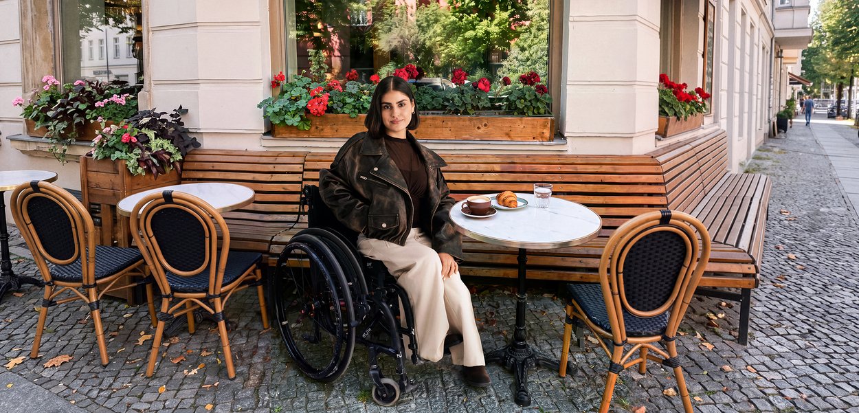 Woman in a wheelchair sitting at an outdoor cafe table with a croissant and coffee, surrounded by empty chairs and flowers.
