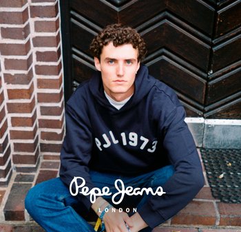 Sudadera azul marino con letras blancas "PJL1973," combinada con jeans azules. El sujeto tiene el cabello rizado y está sentado cerca de una pared de ladrillos texturizada.