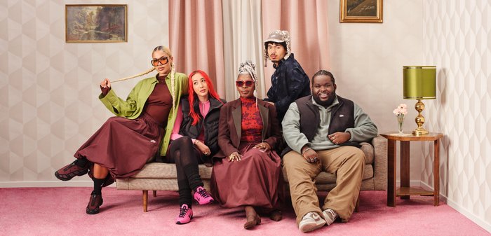 Cinco personas están sentadas en un sofá beige, vistiendo atuendos coloridos. Entre los detalles destacados se encuentran una chaqueta verde, faldas burdeos y suéteres con estampados.