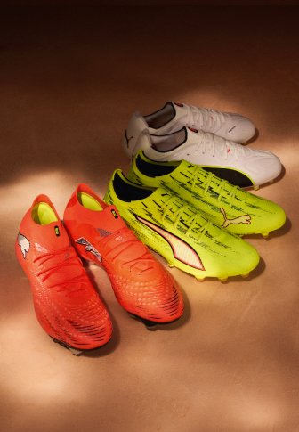 Tri para nogometnih čevljev Puma v rdeči, neonsko rumeni in beli barvi, postavljeni ena ob drugo na rjavi površini.