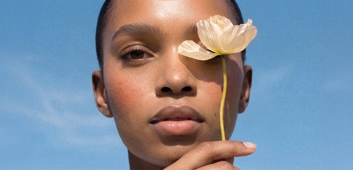 Femme à la peau lisse tenant une fleur pâle qui couvre un œil devant un ciel bleu clair.